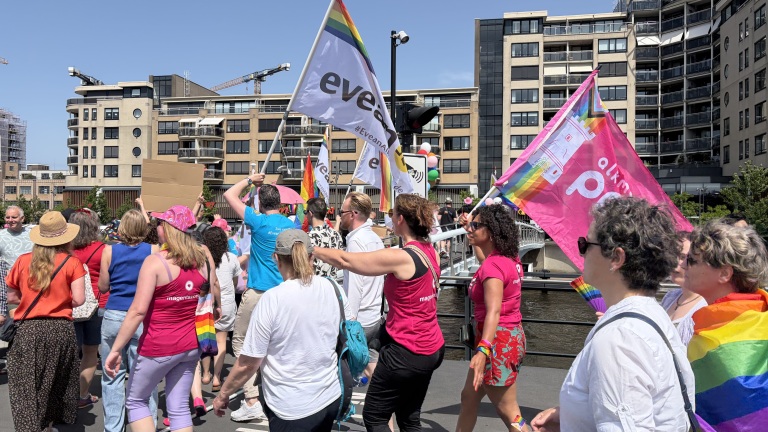 Groep mensen loopt in een parade met regenboogvlaggen en kleurrijke kleding, omringd door stedelijke gebouwen.