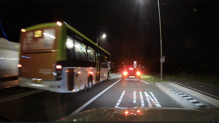 Een stadsbus en een auto rijden 's nachts op een verlichte weg met wegmarkeringen en een bushalte.