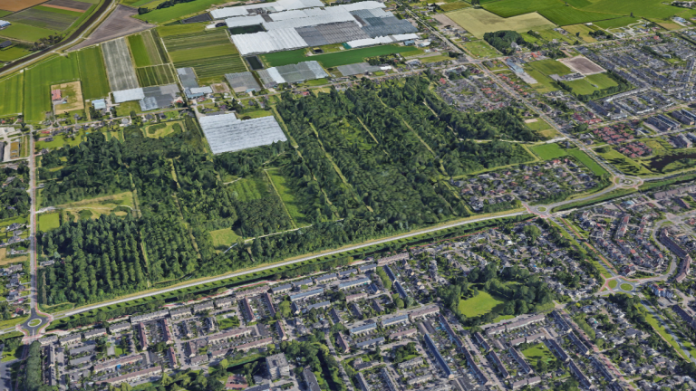 Luchtfoto van een stedelijk gebied met woonwijken, groene bossen en kassen omgeven door landbouwvelden.