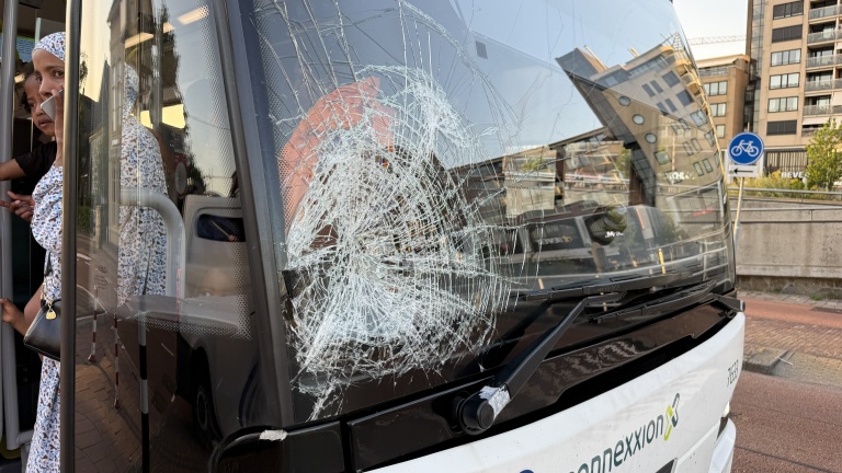 Bus met gebarsten voorruit, vrouw en kind bij de deur in stedelijke omgeving.