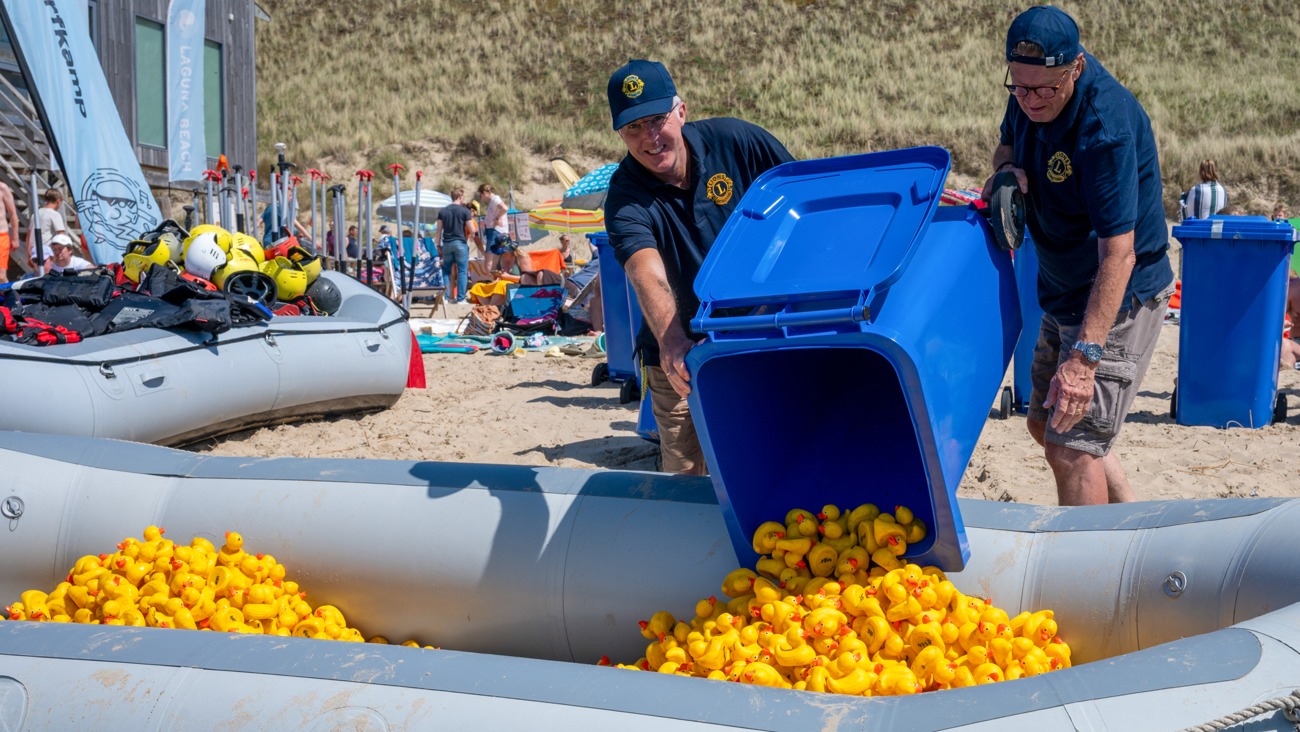 Twee mannen legen een blauwe container vol gele rubberen eendjes in een opblaasboot op een strand.