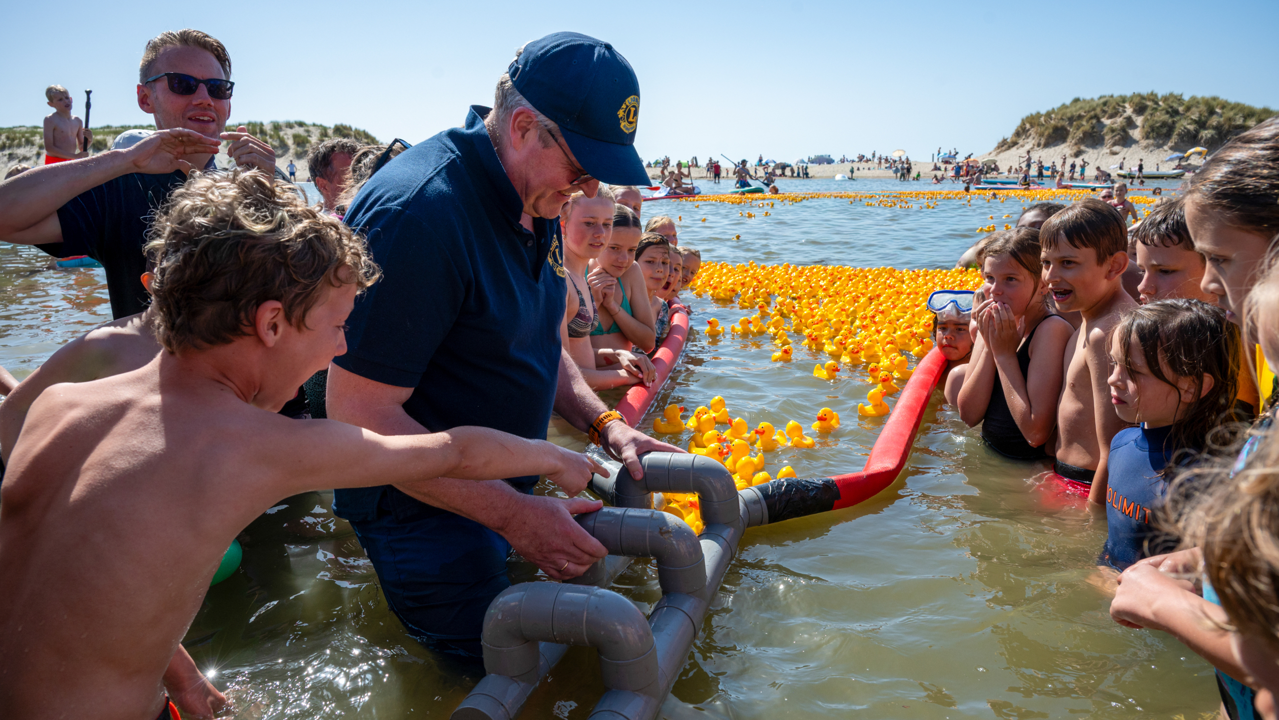 Een groep kinderen kijkt naar een man in het water die een constructie vasthoudt omringd door gele rubberen eendjes.