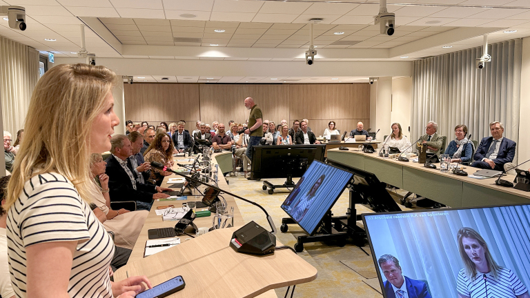 Vrouw spreekt voor een volle vergaderzaal, met schermen die haar toespraak en een tekst over een afscheid laten zien.
