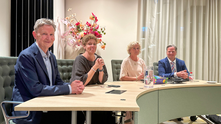 Vier mensen zitten aan een tafel in een vergaderruimte, met bloemen op de achtergrond.