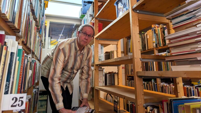 Man in een gestreept overhemd zoekt boeken in een bibliotheek tussen houten planken vol boeken.