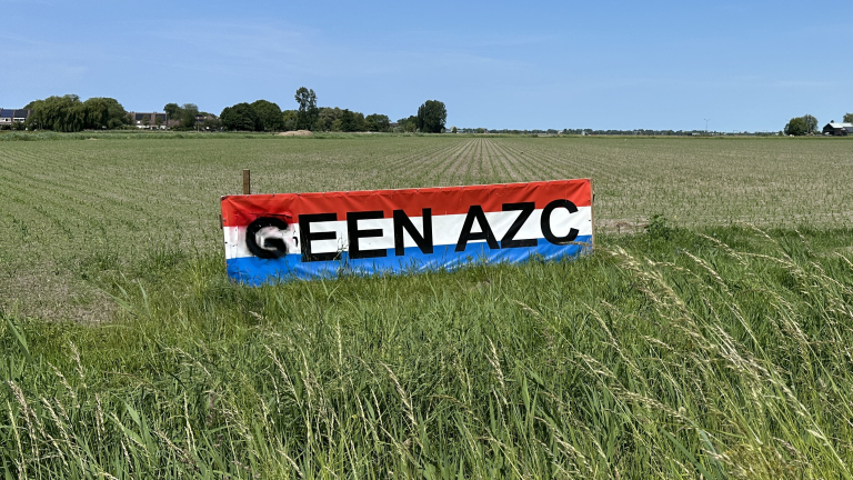 Spandoek met tekst GEEN AZC in rood-wit-blauw bij een akker, met boerderijen en bomen op de achtergrond