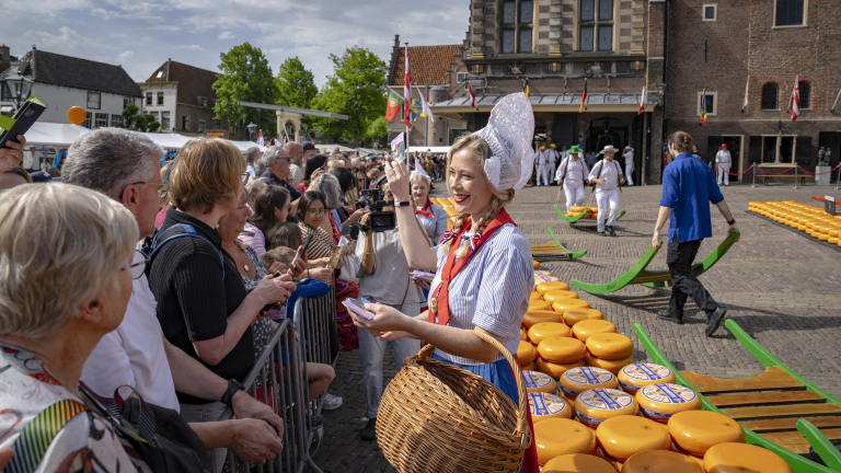 Vrouw in traditionele klederdracht deelt flyers uit aan een menigte bij een kaasmarkt met stapels kaas op de achtergrond.