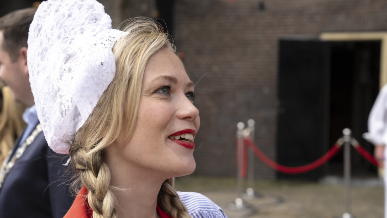 Vrouw met vlechten en een wit kanten kapje kijkt naar links, met achtergrond van een bakstenen muur en rode koorden.