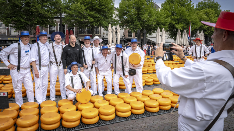 Mensen in witte kleding poseren met grote gele kazen op een markt, terwijl een man een foto van hen maakt.