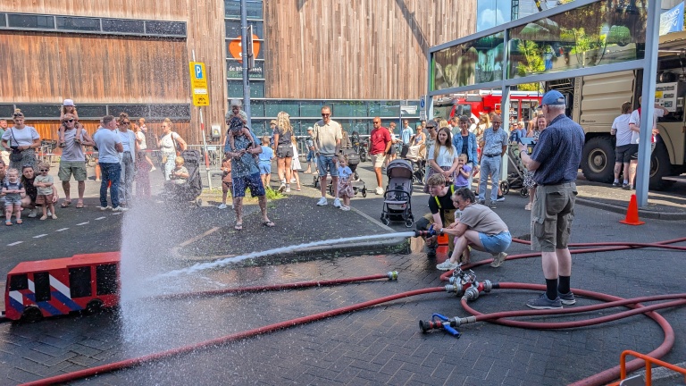 Kinderen spuiten water met een brandslang, omringd door toeschouwers op een zonnige dag bij de brandweer.