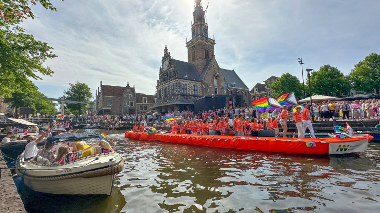 Drukke grachtenparade met mensen in oranje shirts op een boot, omringd door regenboogvlaggen en toeschouwers langs de kade.