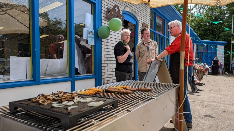 Een barbecue met gegrild vlees en uien staat voor een gebouw met ballonnen, waar mensen samen praten.