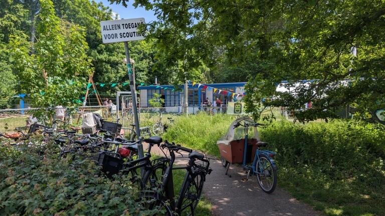 Fietsen geparkeerd voor een scoutingsgebouw met kleurrijke vlaggen, omringd door groen. Een bord vermeldt "Alleen toegang voor scouting".