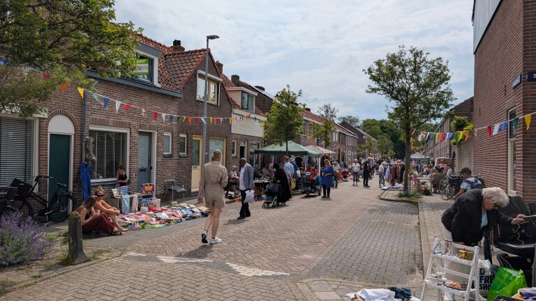 Straatmarkt met mensen die langs kraampjes lopen en tweedehands spullen bekijken, kleurrijke vlaggetjes hangen boven de straat.