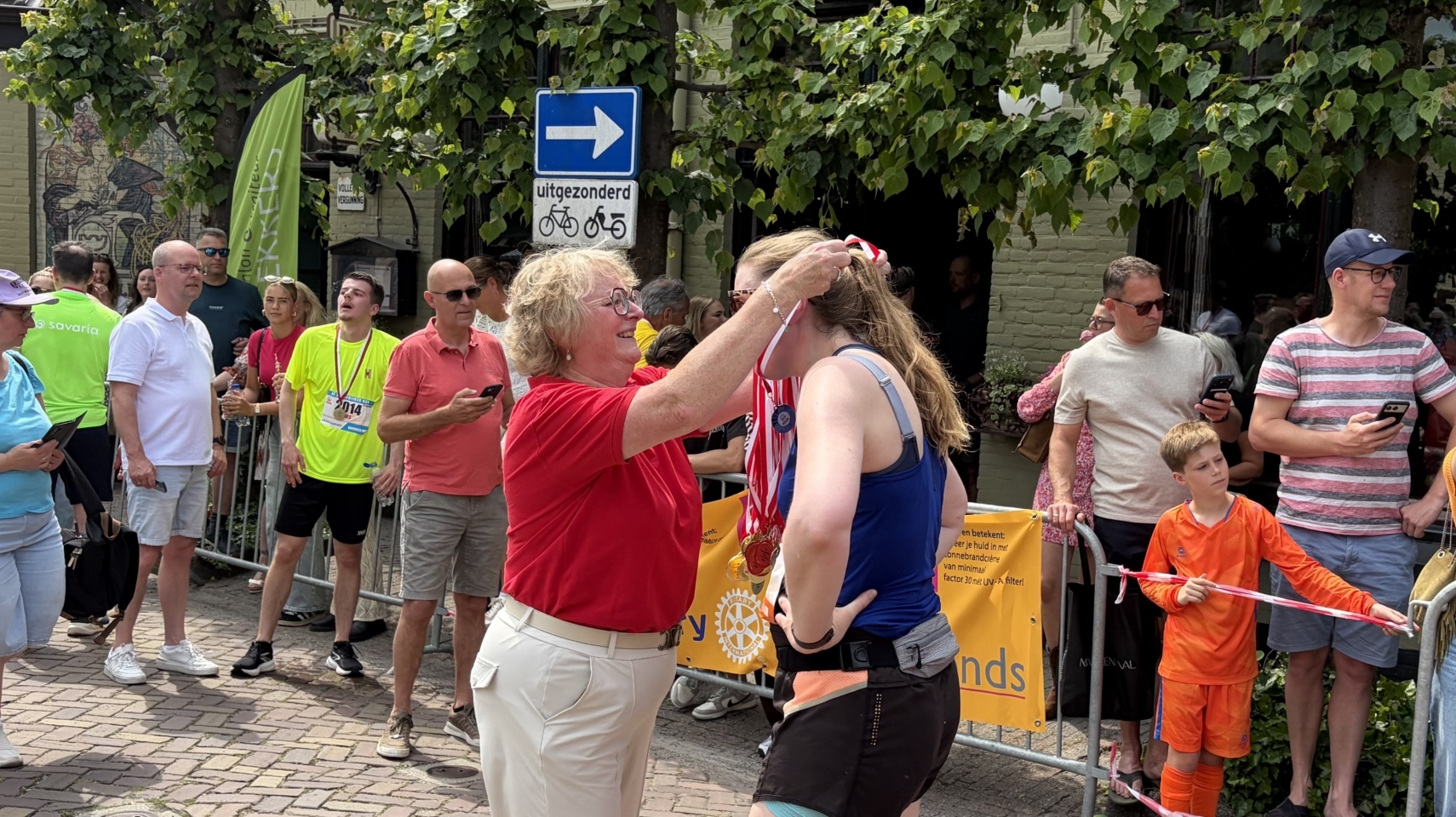 Een vrouw hangt een medaille om de nek van een hardloper terwijl toeschouwers toekijken langs de route.