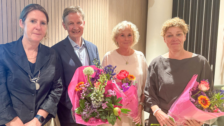 Vier mensen poseren samen, waarvan twee vrouwen bloemenboeketten vasthouden, in een formele setting.