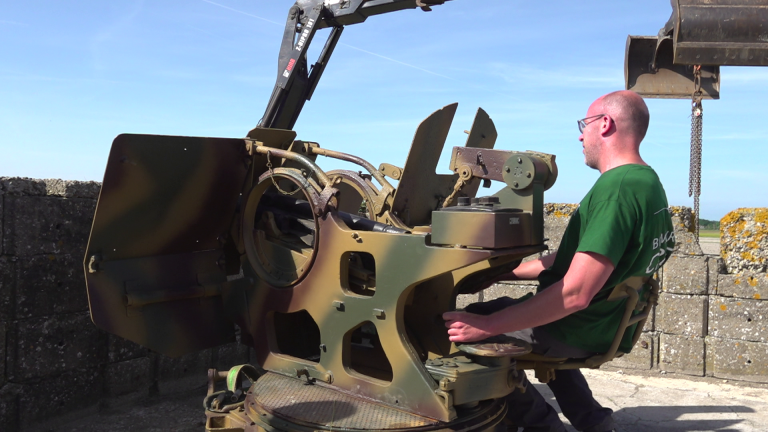 Man zittend op een oude militaire geschutsstelling, met een groene T-shirt en een heldere lucht op de achtergrond.