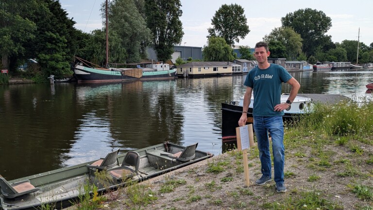 Een man in een T-shirt staat aan de waterkant, naast een bordje, met boten en groene bomen op de achtergrond.