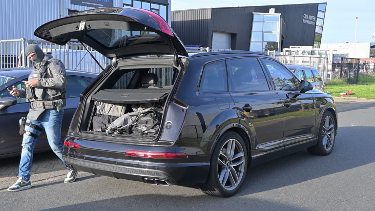 Een politieagent in tactische uitrusting staat naast een geopende kofferbak van een zwarte SUV op een industrieel terrein.