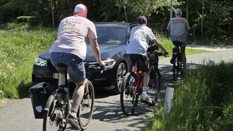 Drie mensen fietsen op een smal pad terwijl er een auto wacht.