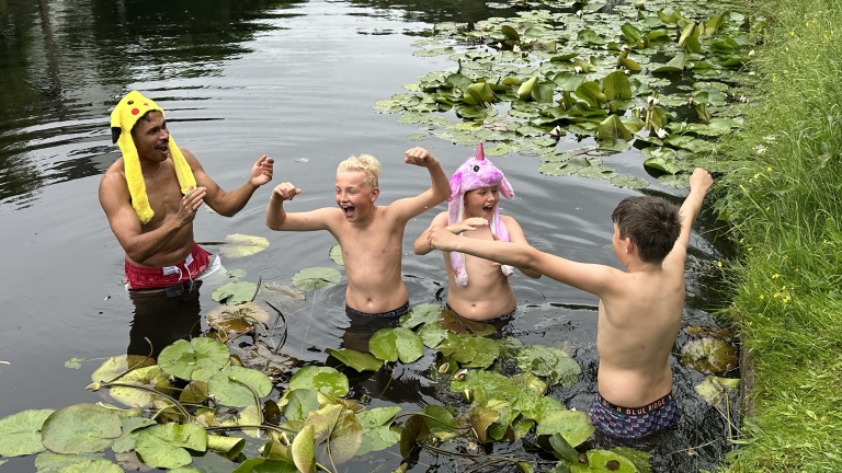 Vier jongens, twee met grappige mutsen, staan lachend in een vijver omringd door waterlelies en planten.