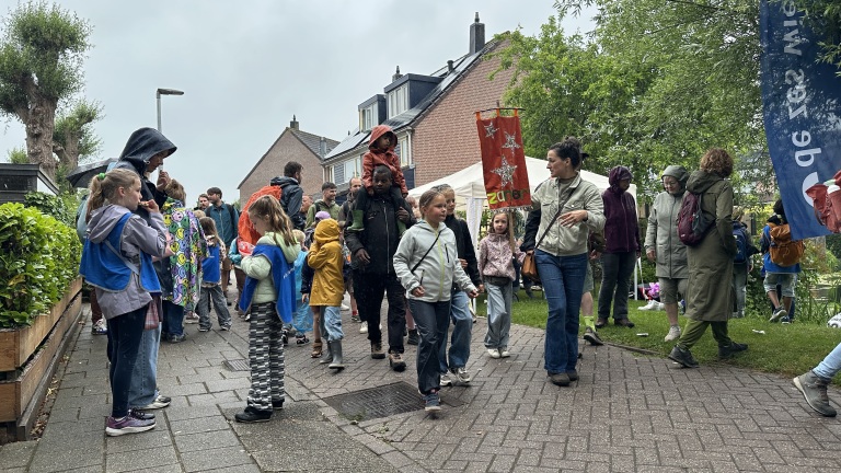 Groep mensen, waaronder kinderen met gekleurde regenjassen, loopt op een regenachtige dag in een woonwijk met groene omgeving.