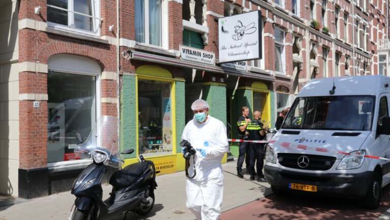Een forensisch onderzoeker in witte kleding en masker loopt langs een afgezette straat bij een winkel, met politie en busje aanwezig.