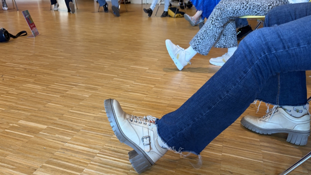 Personen zitten in een kring op stoelen, met de focus op een uitgestoken been in jeans en beige laarzen.