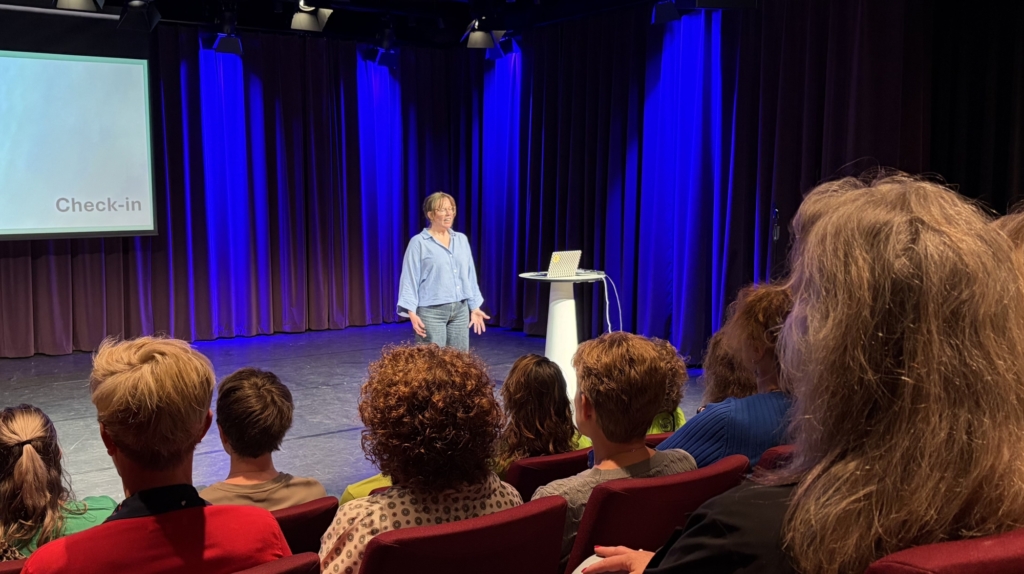 Een persoon geeft een presentatie op een podium met blauwe achtergrondverlichting, terwijl een publiek toehoort.