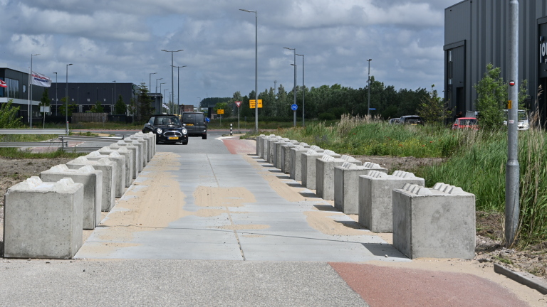 Een weg met betonnen blokken aan beide zijden, een zwarte auto nadert, industriële gebouwen en groen op de achtergrond.