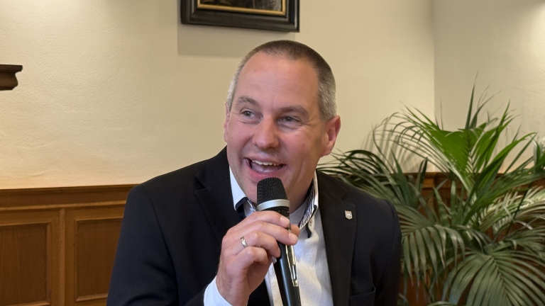 Man in een pak spreekt in een microfoon met een glimlach, op de achtergrond staan planten en houten lambrisering.