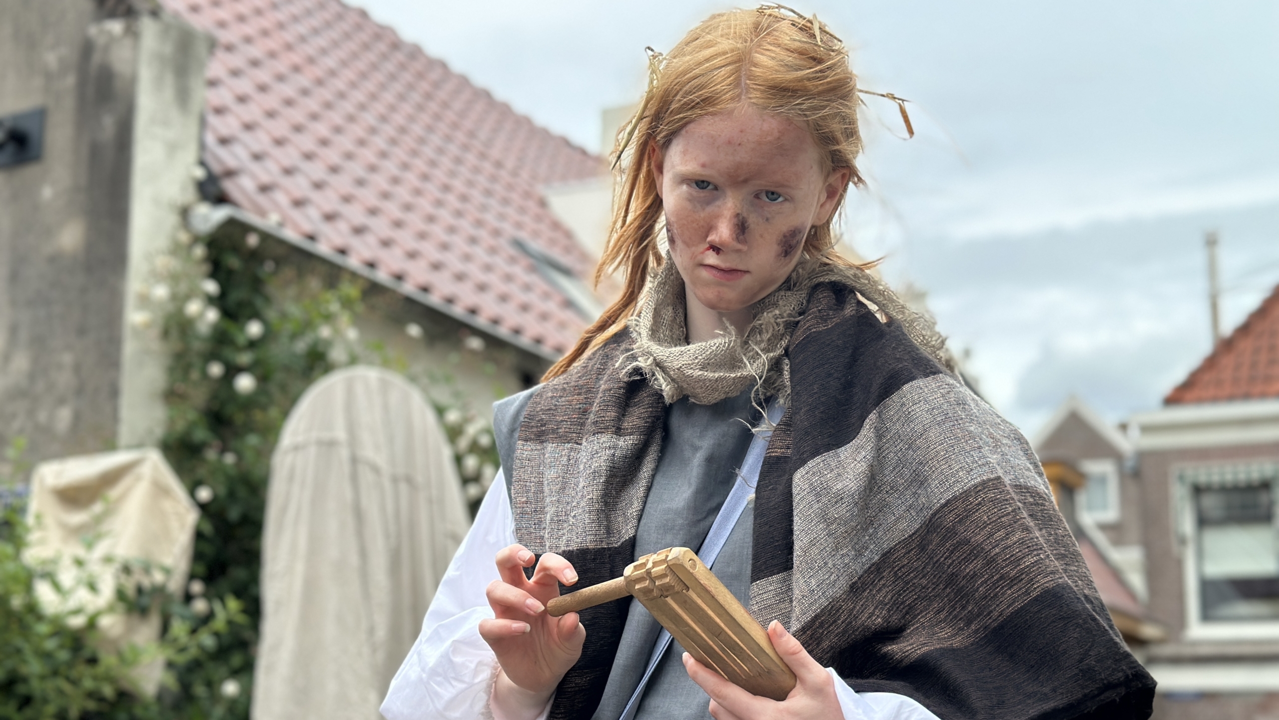 Persoon in een historische outfit met een verweerd gezicht, houdend een houten voorwerp in een buitenomgeving.