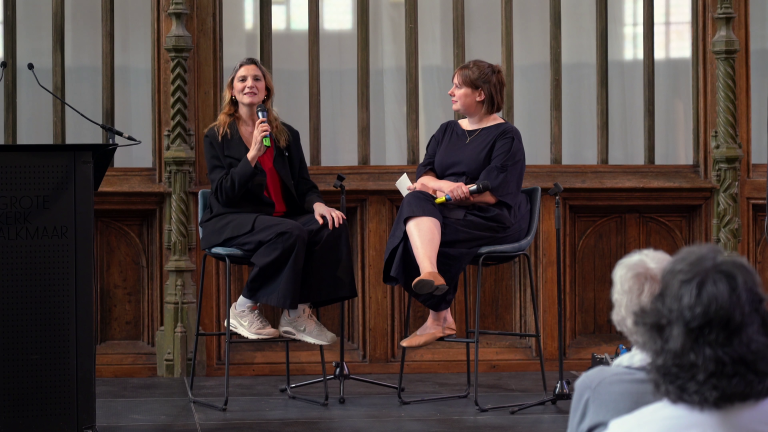 Twee vrouwen zitten op een podium met microfoons, terwijl ze een gesprek voeren voor een publiek in een historische zaal.