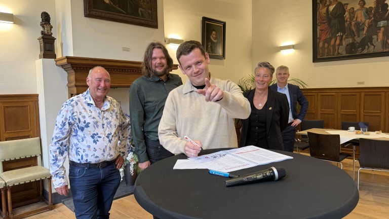 Een groep van vijf mensen staat rond een tafel; in het midden ondertekent iemand een document, met een microfoon op de voorgrond.