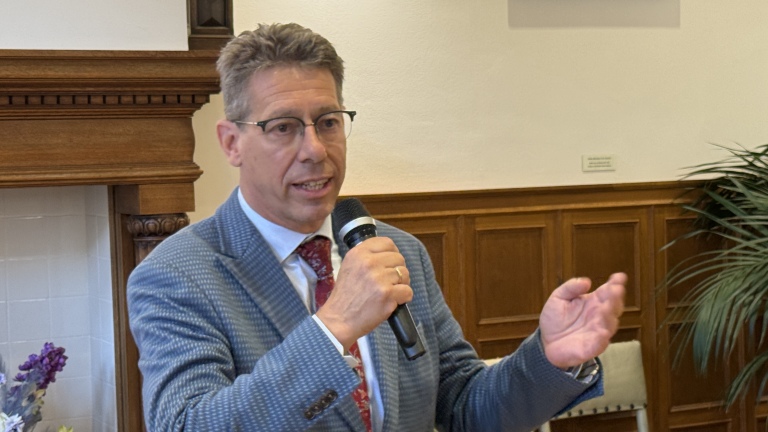 Man in een blauw pak met bril en stropdas spreekt in een microfoon met een handgebaar, staand in een kamer met houten lambrisering.