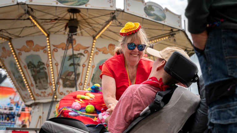 Vrouw met zonnebril en feesthoed lacht naar een persoon in een rolstoel bij een draaimolen.