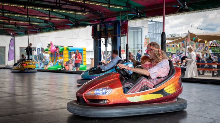 Een kind en een volwassene lachen samen in een botsauto op een kermis, omgeven door andere attracties en mensen.