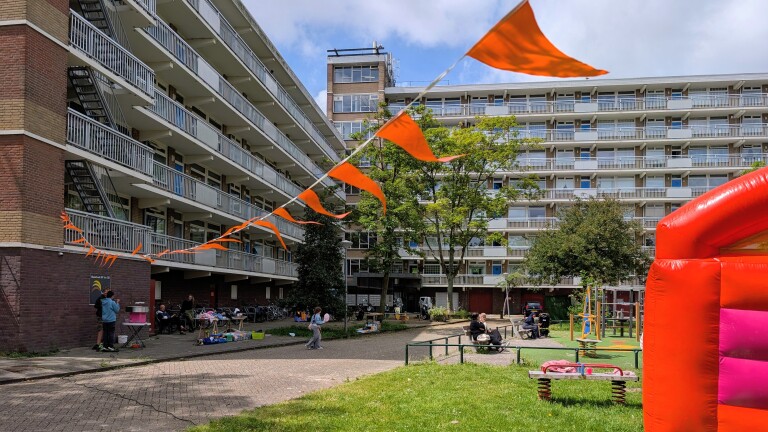 Flatgebouw met balkons en oranje vlaggetjes, mensen op een plein en een felgekleurde speelstructuur op de voorgrond.