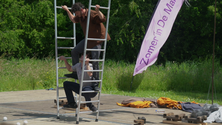 Twee mensen balanceren op een metalen ladder op een buitenpodium, naast een vlag met de tekst "Zomer in De Marne."