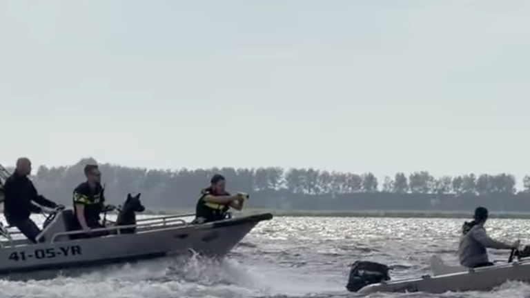 Een politieboot achtervolgt een kleiner vaartuig op het water, met agenten en een hond aan boord.