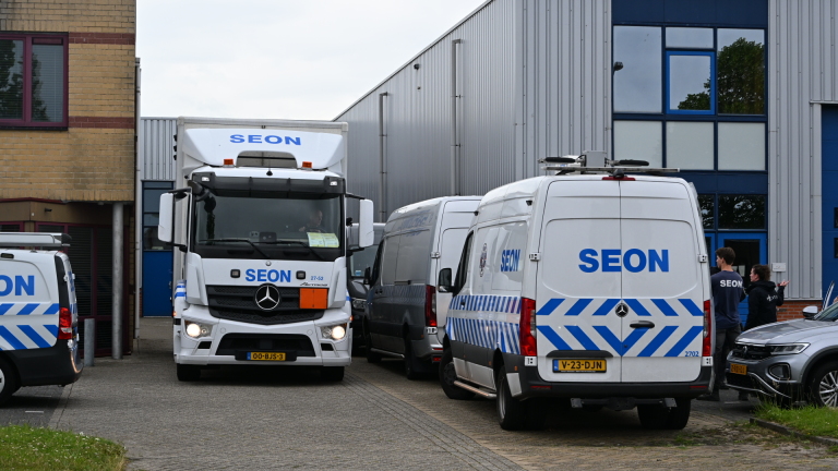 Vrachtwagens en bestelwagens van SEON staan geparkeerd voor een grijs industrieel gebouw, omringd door mensen.