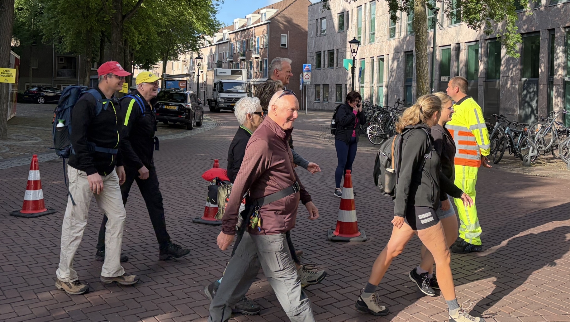 Mensen lopen over een straat met bomen en bakstenen gebouwen. Sommigen dragen rugzakken en wandelkleding.
