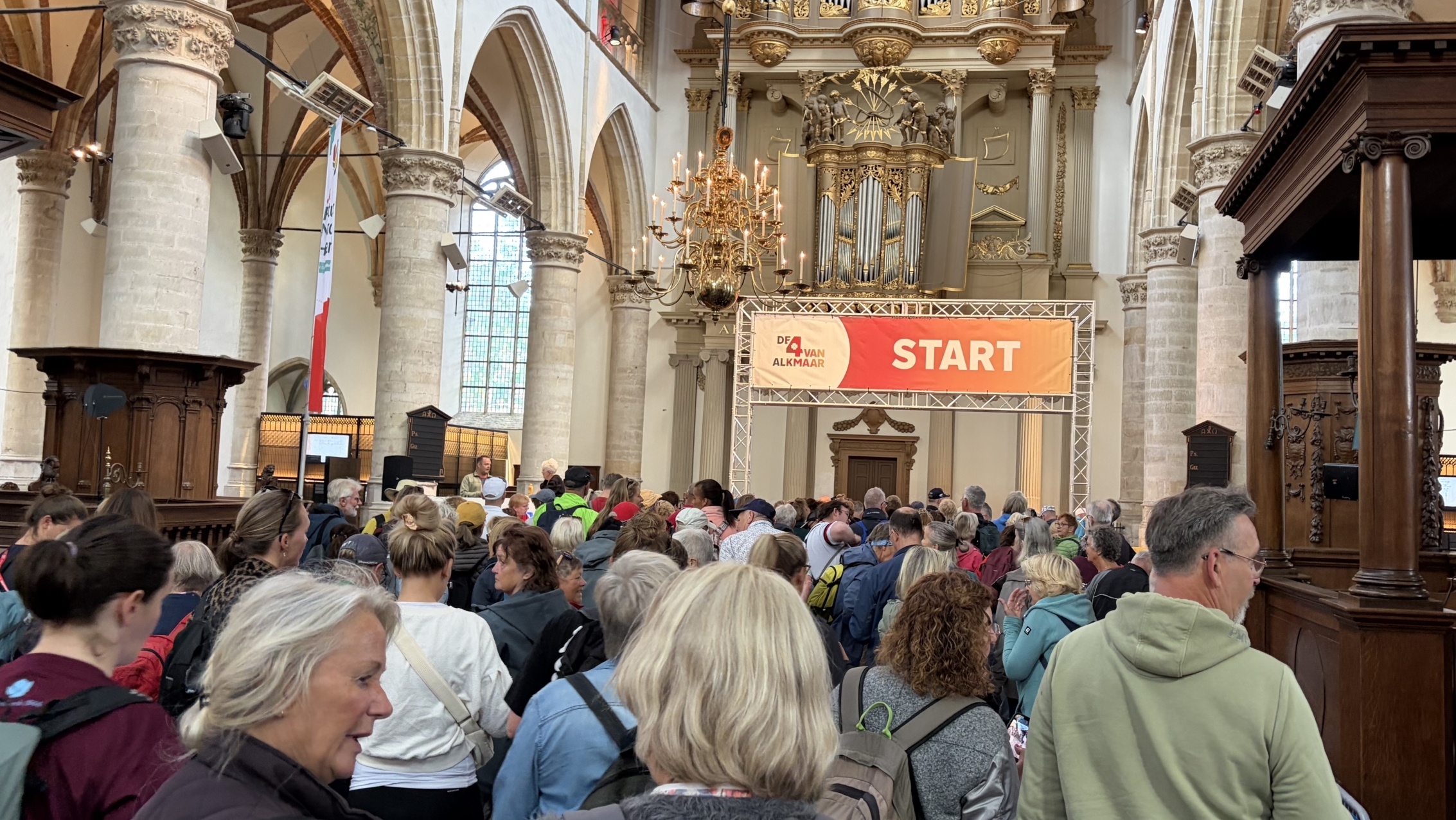 Een groep mensen staat in een kerk bij een bord met "START", vermoedelijk voor een evenement met de naam "De 4 Van Alkmaar".