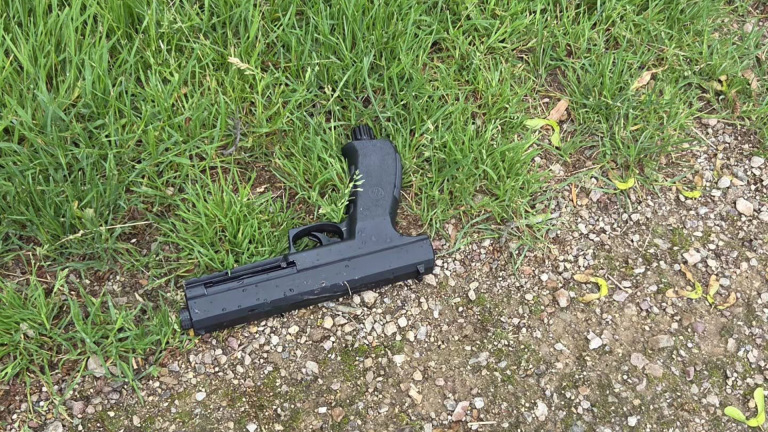 Een zwart pistool ligt op de grond, deels op gras en deels op een grindpad.