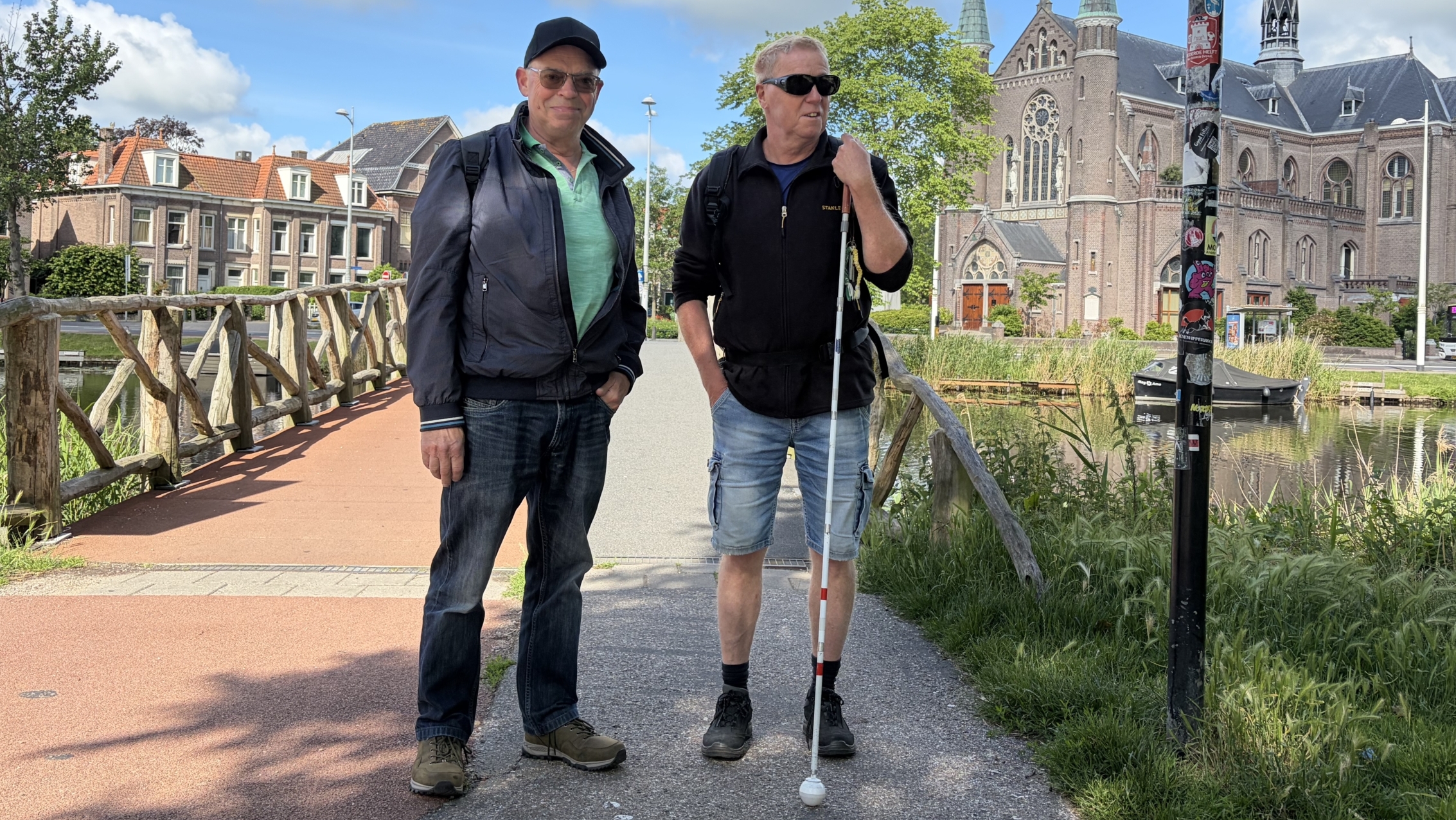 Twee mannen staan op een brug met een kerk op de achtergrond; de man rechts heeft een taststok.