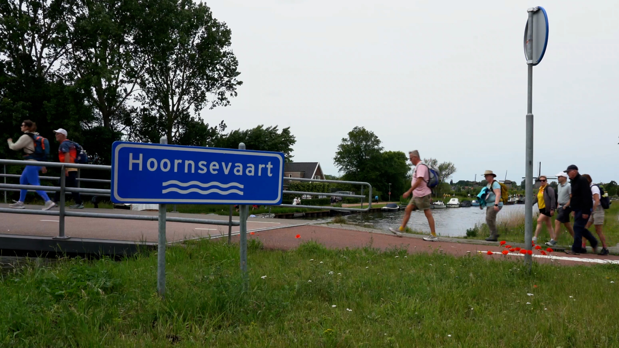 Wandelaars lopen langs een weg bij een bord met de tekst "Hoornsevaart", omgeven door gras en bomen.