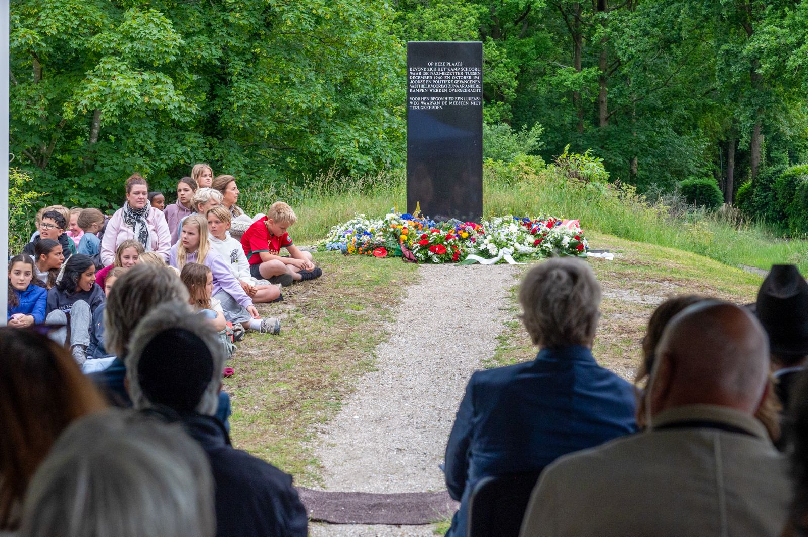 Mensen, waaronder kinderen, zitten voor een gedenksteen omringd door bloemen in een groene omgeving.