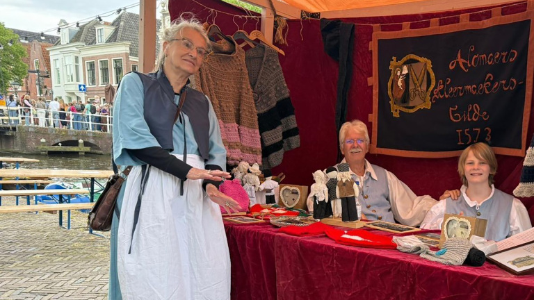 Drie mensen in historische kledij bij een kraam van het Alkmaers Kleermaeckers Gilde, met handwerkproducten en een spandoek.