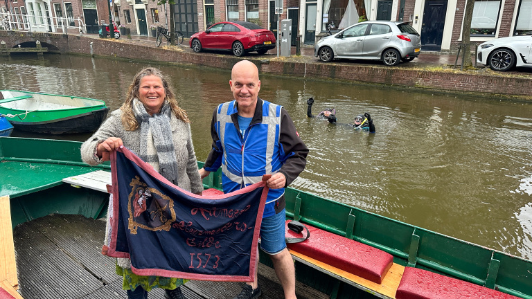 Twee mensen houden een vlag vast op een boot in een kanaal, met duikers zichtbaar in het water op de achtergrond.