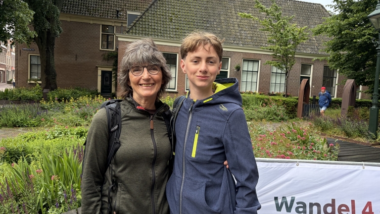 Een vrouw en een jongen glimlachen samen buiten voor een huis met een tuin vol bloemen, een spandoek met tekst is deels zichtbaar.